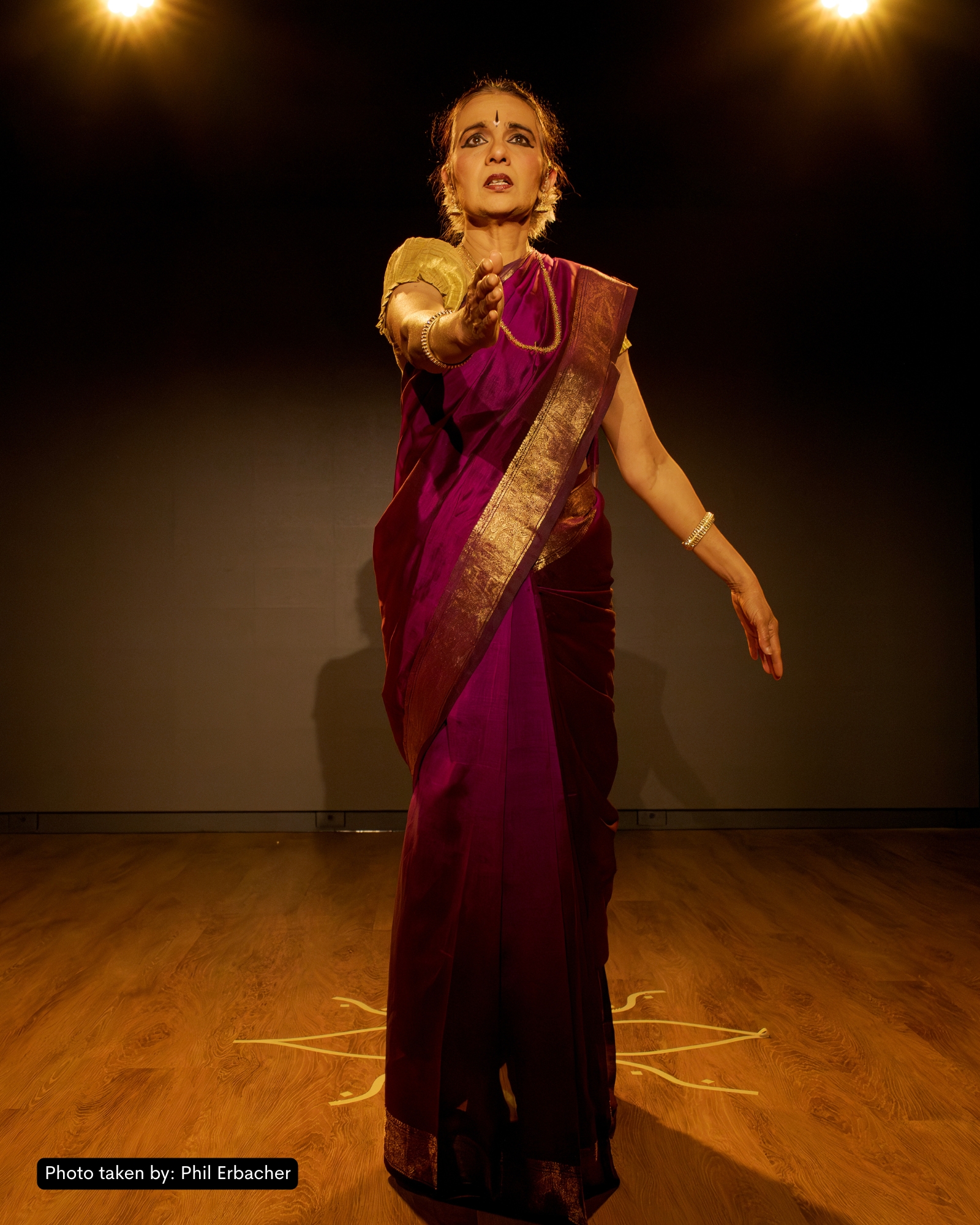 Production still from And What Will People Say? — Kersherka mid-Bharatanatyam pose, lit warm amber against a deep-shadowed stage.